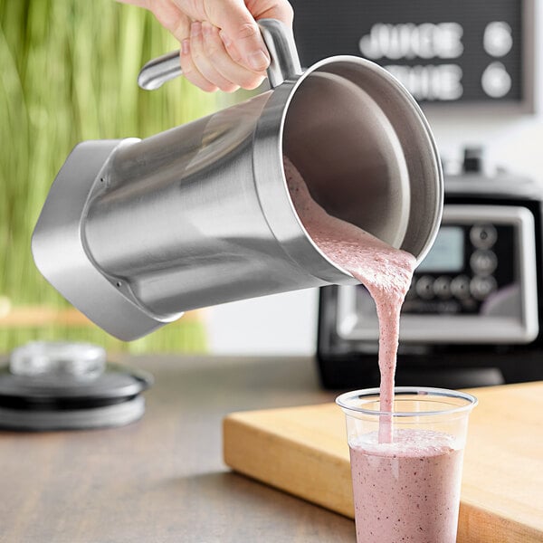 A hand pours a pink smoothie into a stainless steel AvaMix blender jar.