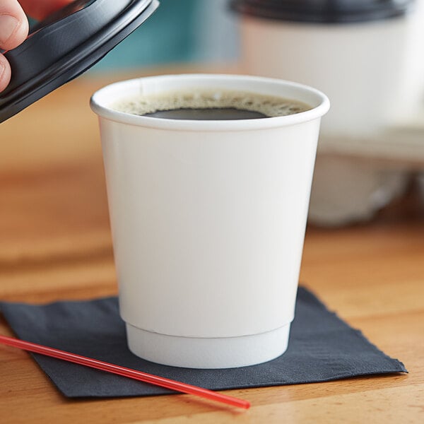 A hand holding a white double wall paper hot cup of coffee with a black lid.