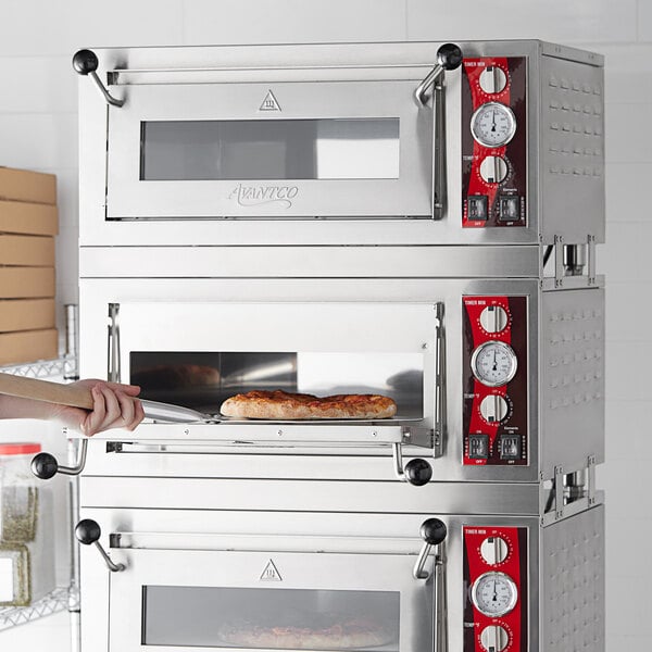 A woman using a wooden stick to put a pizza in an Avantco triple deck pizza oven.