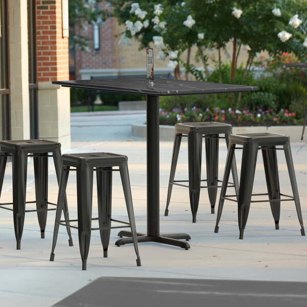 A Lancaster Table & Seating rectangular counter height table with a grey surface and a white stripe on a patio with black stools.