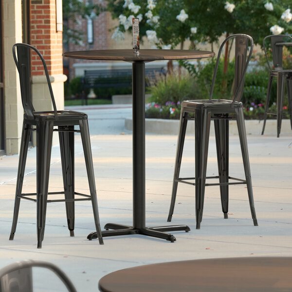 A Lancaster Table & Seating round bar height table with a textured finish and cross base plate on a patio with three black metal chairs.