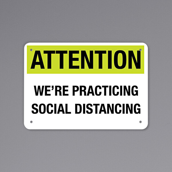 A black and yellow aluminum sign that says "Attention / We're Practicing Social Distancing"