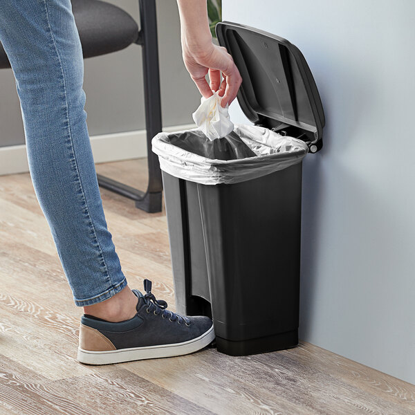 A hand putting a white bag in a Lavex black rectangular step-on trash can.