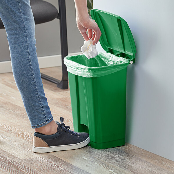 A person wearing jeans and shoes steps on a green Lavex rectangular step-on trash can to throw a white bag inside.