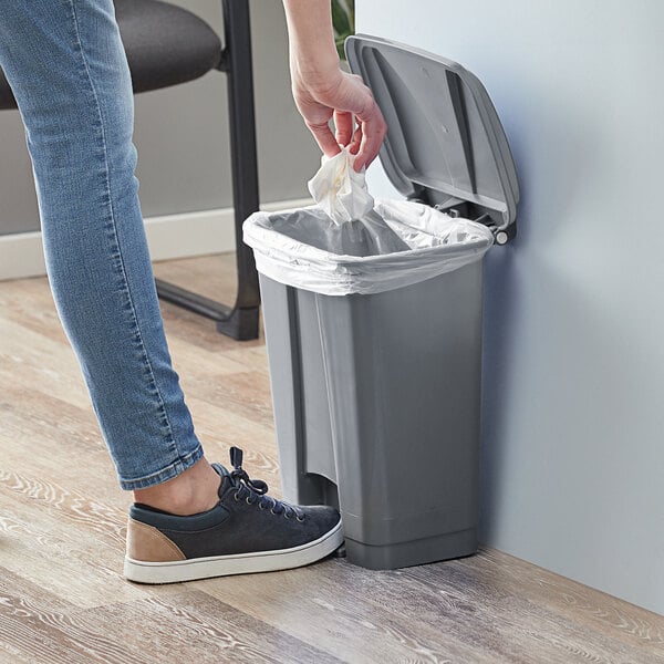 A person putting a white bag in a Lavex gray rectangular step-on trash can.