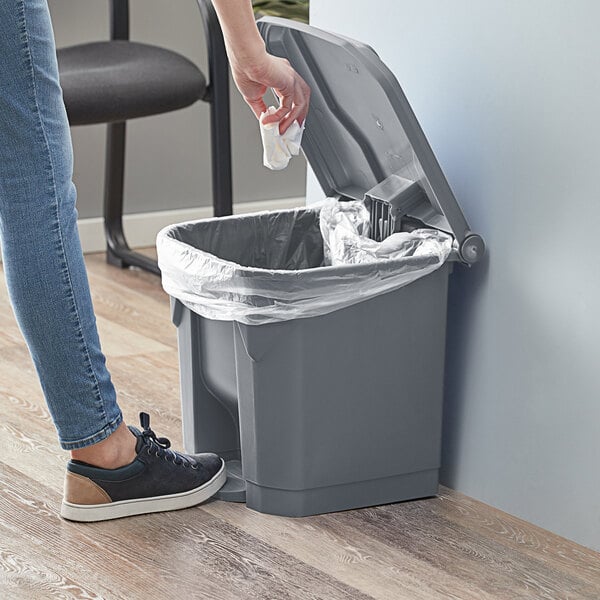 A person in jeans using their foot to open a Lavex rectangular step-on trash can to throw a white cloth inside.