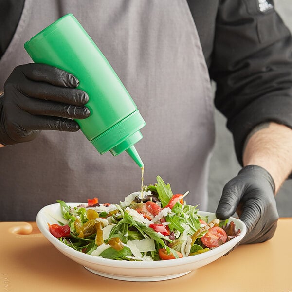A person in a black glove using a Choice green wide mouth squeeze bottle to pour sauce on a bowl of salad.
