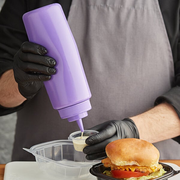 A person in gloves pouring sauce from a purple Choice wide mouth squeeze bottle onto a burger.