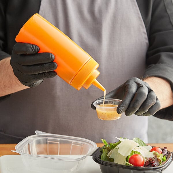 A person in black gloves using a Choice orange squeeze bottle to pour sauce into a small container.