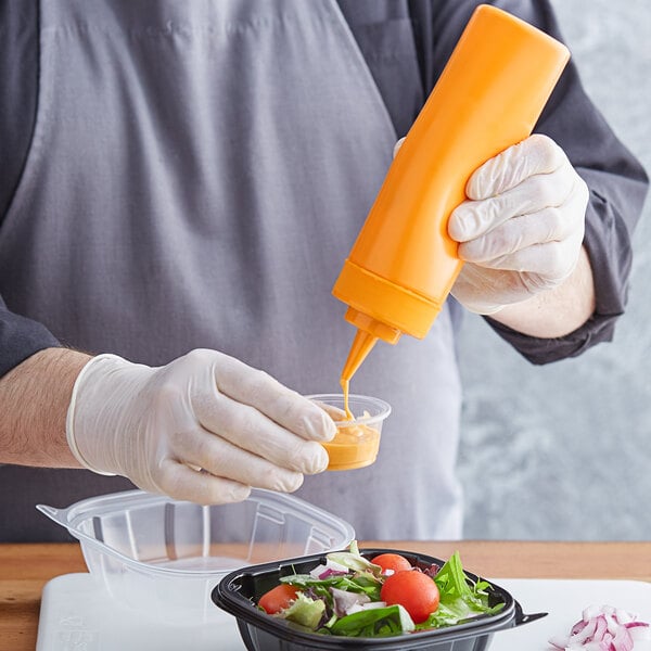 A person in a white apron using a Choice orange squeeze bottle to pour yellow liquid onto a salad in a plastic container.