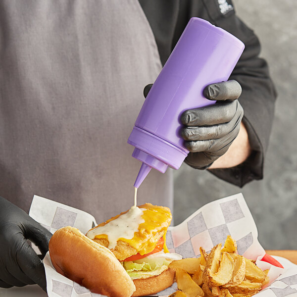A person in gloves using a Choice purple wide mouth squeeze bottle to pour sauce on a burger.