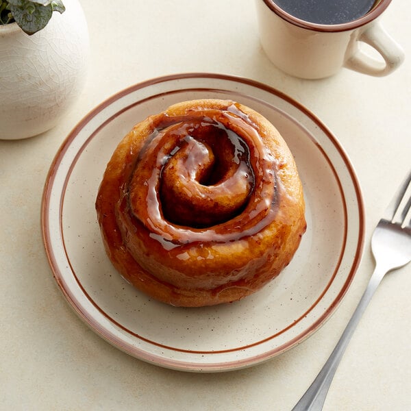 A cinnamon roll on an Acopa brown speckle narrow rim stoneware plate with a fork next to a cup of coffee.