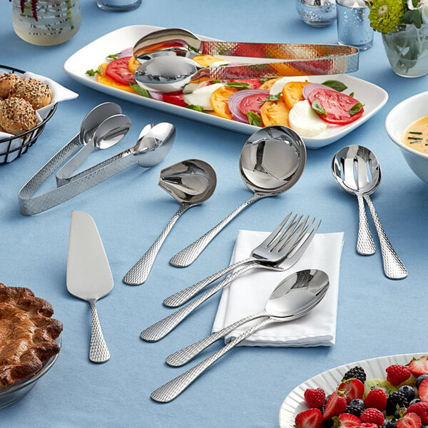 A table set with Acopa Industry 18/8 stainless steel serving utensils, including spoons, forks, and tongs, with a variety of food.