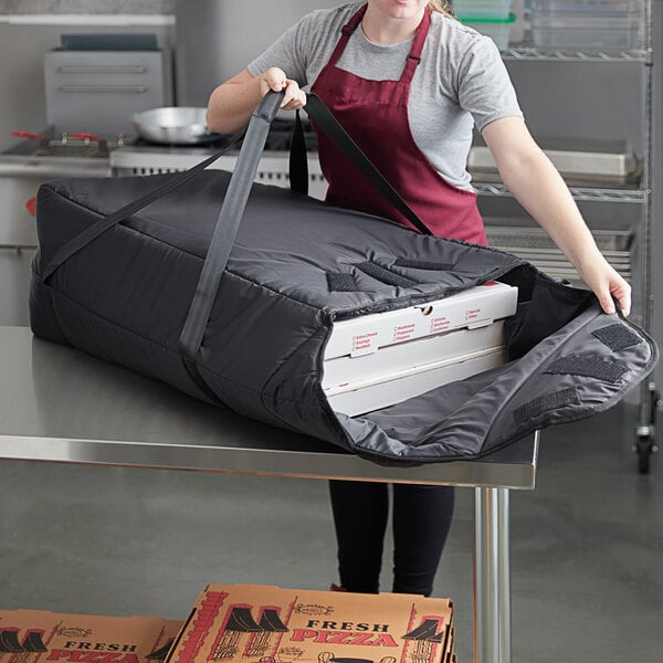 A woman in a red apron holding a large black Vesture NextPhase insulated pizza delivery bag with a pizza inside.