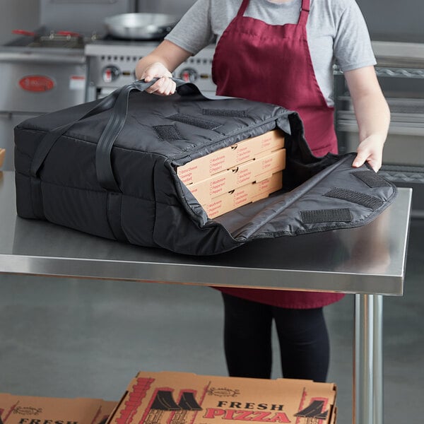 A woman in a red apron holding a Vesture NextPhase black insulated pizza delivery bag containing a pizza.