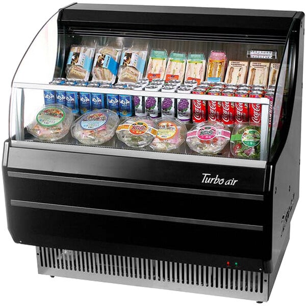 A black Turbo Air horizontal air curtain display case with food inside including salad in round plastic containers with labels.