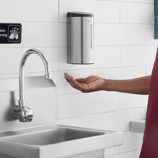 A person using a Lavex Stainless Steel automatic liquid soap / sanitizer dispenser to wash their hands in a public bathroom.