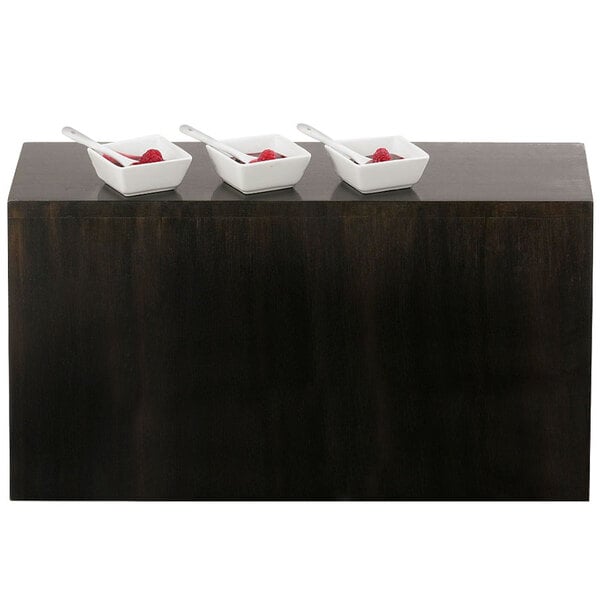 A row of white bowls with spoons on top of a black Cal-Mil Midnight plate riser.
