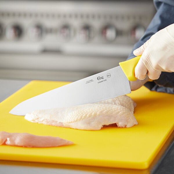 A person using a Choice 10" Chef Knife with a yellow handle to cut chicken on a yellow cutting board.
