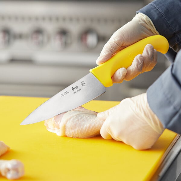A person holding a Choice 6" Chef Knife with a yellow handle and cutting a chicken.