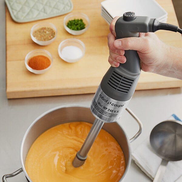 A hand using a Robot Coupe Mini Immersion Blender to mix a bowl of food on a counter.