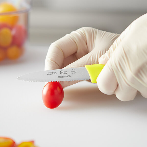 A person in gloves uses a Choice serrated paring knife with a neon yellow handle to cut a tomato.