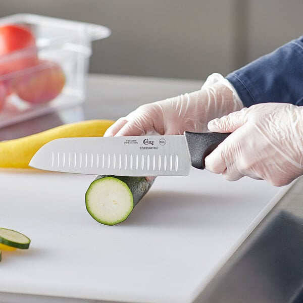 A person in a white coat using a Choice Santoku knife to cut cucumbers on a counter in a professional kitchen.