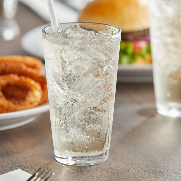A Pasabahce beverage glass filled with ice water and a straw on a table with a burger.
