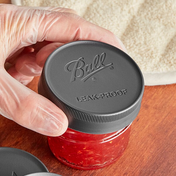 A hand holding a Ball Regular Mouth Canning Jar filled with red liquid with a Ball black plastic lid.