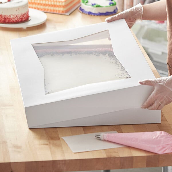 A person holding a Baker's Mark white bakery box with a clear window over a cake.