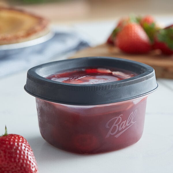 A Ball plastic freezer jar filled with red strawberry jam on a table.