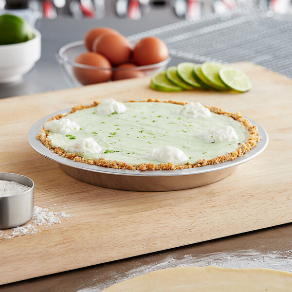 A pie with lime and whipped cream in a Choice 9" x 1 1/4" 21 Gauge Aluminum Pie Pan on a wooden cutting board.