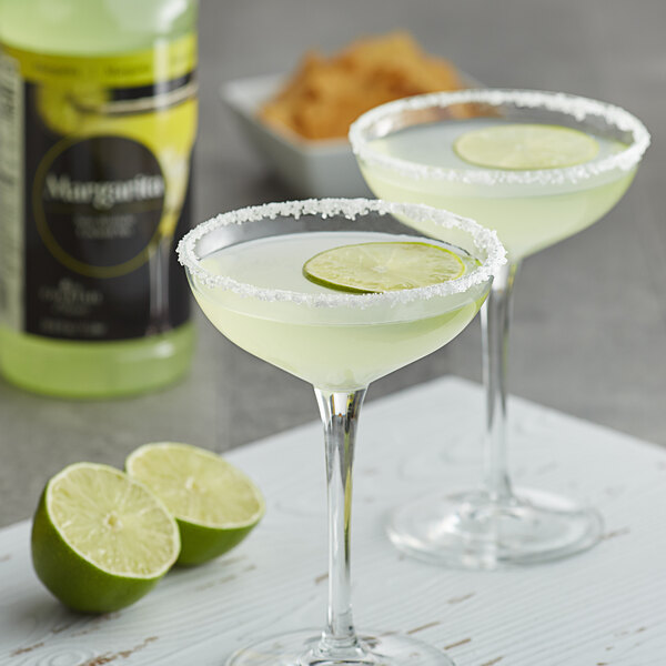 A pair of glasses of Regal Cocktail Margarita with lime slices and a bottle on a table.