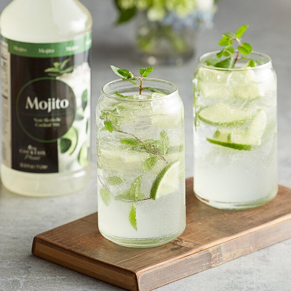 A bottle of Regal Cocktail Mojito Mix on a wooden board with a glass of mojito and lime slices.