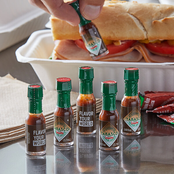 A hand holding a TABASCO® Chipotle Hot Sauce mini bottle next to a sandwich.