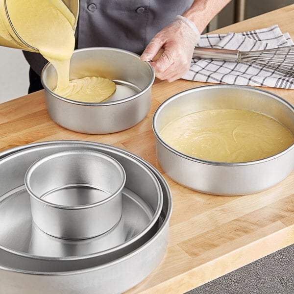 A person pouring yellow cake batter into a round metal cake pan.
