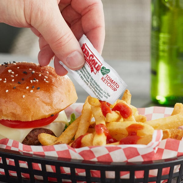 A hand putting a Red Gold ketchup packet on a burger on a table with red and white checkered fabric.
