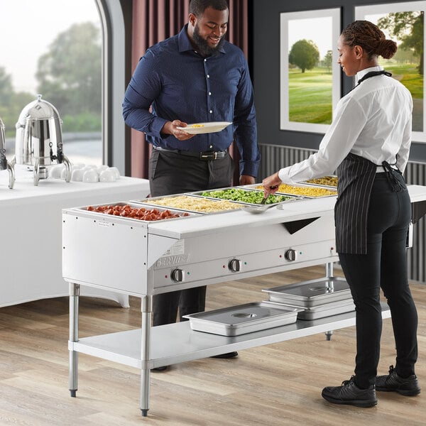 A man and woman standing next to a ServIt electric steam table in a hotel buffet.