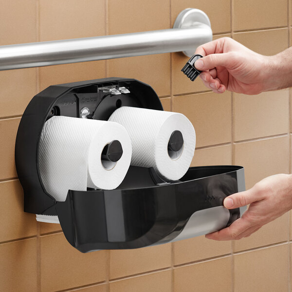 A hand holding a roll of toilet paper over a black San Jamar toilet tissue dispenser.