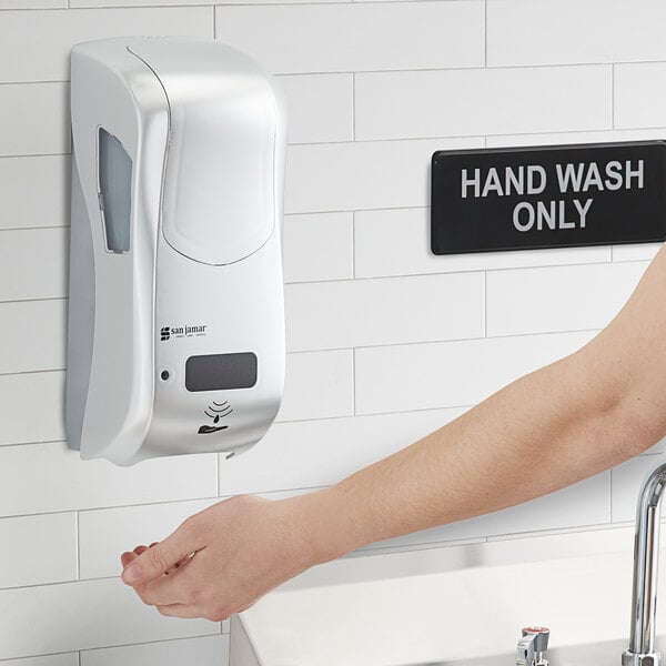 A person using a San Jamar silver hybrid automatic foam hand sanitizer dispenser.