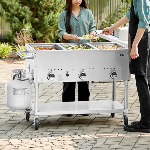 A man and woman serving food from a Backyard Pro steam table in an outdoor setup.