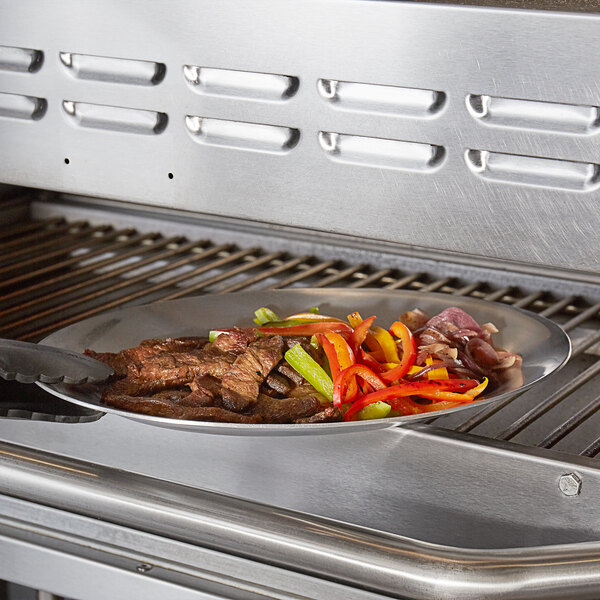 An oval aluminum sizzler platter with food cooking on a grill.
