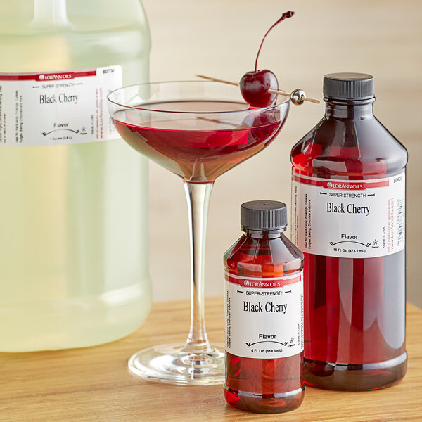 A bottle of LorAnn Oils Black Cherry Flavor next to a glass of red liquid with a cherry on top.