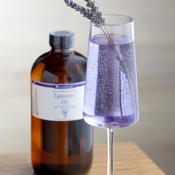 A bottle of LorAnn Oils All-Natural Lavender Super Strength Flavor on a table with a glass of lavender drink.