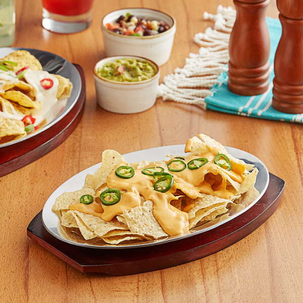 A mahogany finished oval aluminum sizzler platter on a table with nachos, cheese, and jalapenos.