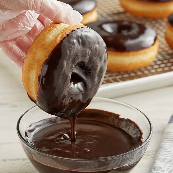 A hand dipping a Rich's Chocolate covered donut into chocolate icing in a bowl.