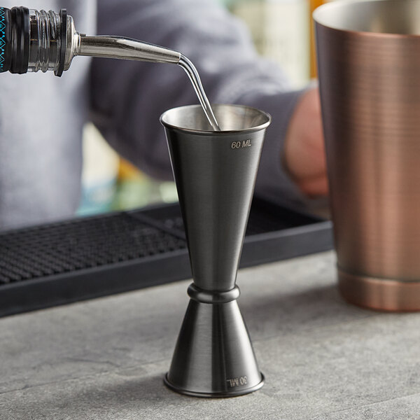 A person using an American Metalcraft black brushed stainless steel Japanese style jigger to pour liquid into a metal cup.