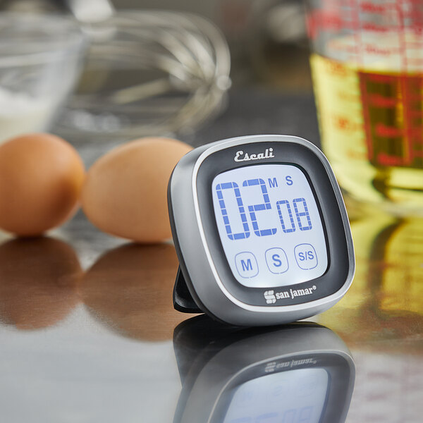 A San Jamar digital kitchen timer on a counter with eggs and a measuring cup.