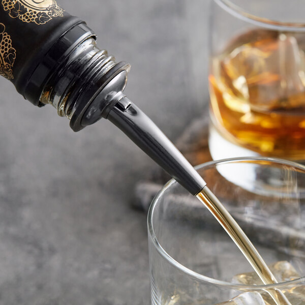 A bottle of American Metalcraft black stainless steel liquor pourer pouring liquid into a glass of whiskey with ice.