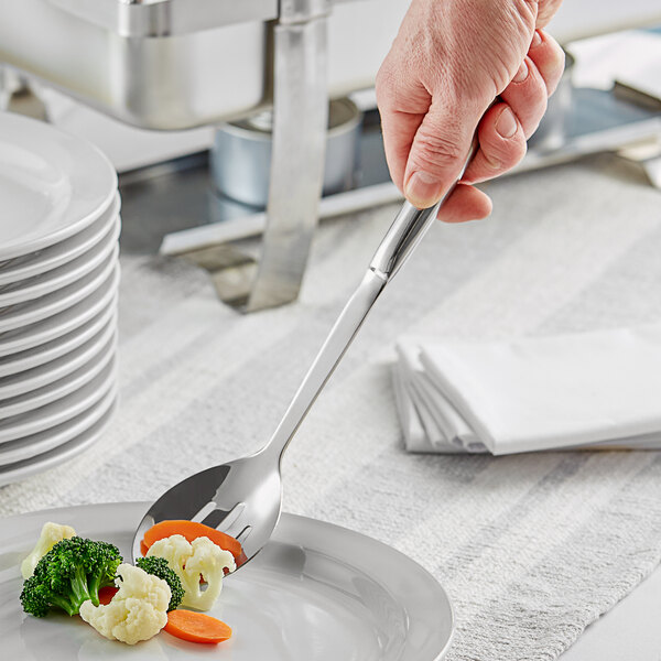 A hand holding a slotted serving spoon over a plate of vegetables.